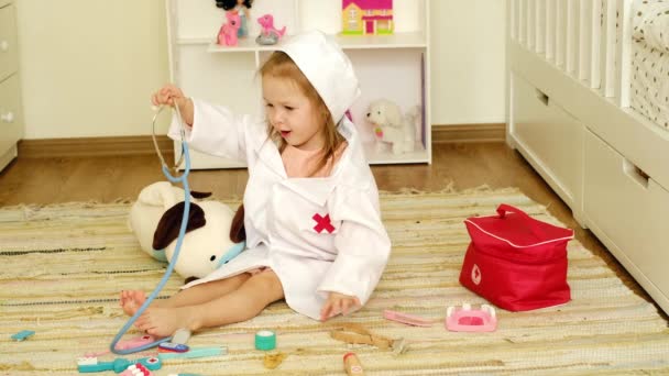 一个两岁的小女孩穿着医疗服扮演医生 为一只带听诊器的玩具狗做手术 可爱的小宝宝一个人在育儿室玩耍 那孩子想成为一名兽医 — 图库视频影像