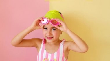 Pembe çizgili mayo giymiş 5 yaşında çekici bir kız, gözlüğünü gözlerine doğru çekiyor. Okul öncesi çocuklar için havuzda yüzme dersleri. Sarı pembe stüdyo arka planı