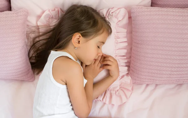 A long-haired girl is sleeping sweetly in a bed with pink pillows, with her hand tucked under her cheek. Pillow with ruffles. Healthy strong children\'s daytime sleep. Baby sleep schedule
