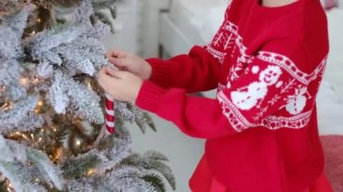Süslü bir süveter içindeki bebek, Noel ağacını yapay karla süsleyen güzel bir köknar ağacının dalına oyuncak bir şeker çubuğu asarak süslüyor. Yılbaşı ve Xmas 'a hazırlanıyorum. Işıklar çelenk