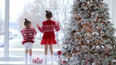 Kızlar cama resim çizer, pervazda durur ve aralık günü büyük pencerenin önünde yağan karı izlerler. Kızların yanındaki ışıklarla süslenmiş Noel ağacı. Yeni yılı bekliyorum. xmas