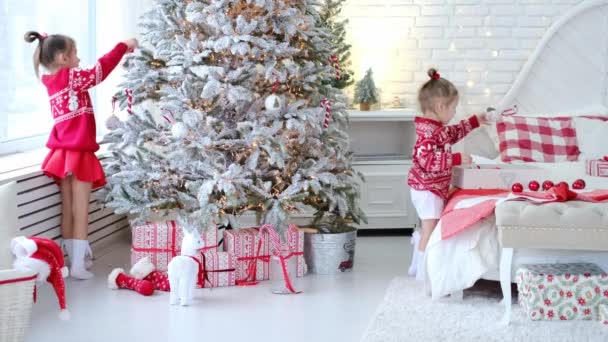 A gyerekek karácsonyi játékokat és golyókat akasztanak egy mesterséges karácsonyfára egy világos fehér szobában. Ajándékok nyaralás csomagolás fekszik a padlón alatt egy lucfenyő. A lányok piros pulcsiba vannak öltözve. karácsony