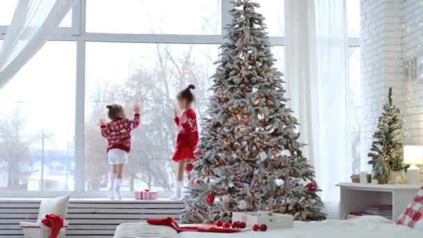 孩子们站在窗台上 在一个有着圣诞树和新年装饰的房间的巨大窗户前 看着12月的第一场雪在外面飘扬 神奇的一天 穿着红色毛衣 头戴装饰品的孩子 — 图库视频影像