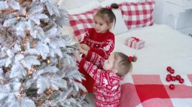 İki tatlı kız kardeş Noel ağacına top asar ve yılbaşı ve Noel için odayı dekore ederler. Yatağın üzerinde kareli yastıklar ve battaniye var. Süslü kırmızı kazaklı çocuklar.