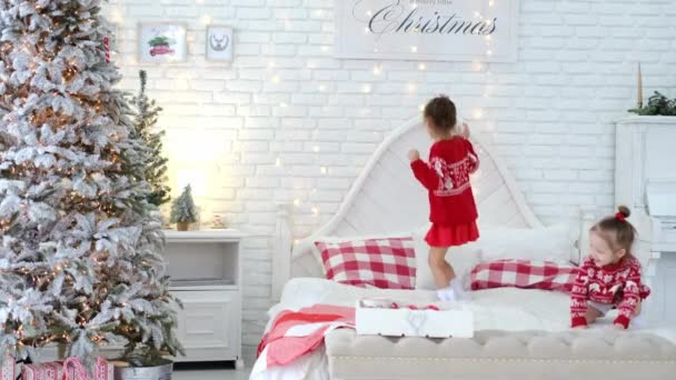 子供たちはジャンプし レンガの白い壁の背景にクリスマスの装飾とクリスマスツリーを備えた親の寝室のベッドでお楽しみください もうすぐ新年が来る 子供のユーモアだ 赤いセーター — ストック動画