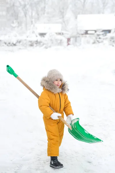 クリーニングと雪の除去のための巨大なプラスチック製の緑のシャベルと黄色のジャンプスーツの子供 雪が辺り一面を覆っていた 雪の霜の冬 女の子は道や歩道から雪を取り除くのに役立ちます — ストック写真