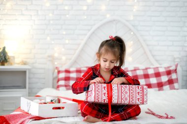 Kareli kırmızı pijamalı bir kız Noel hediyesini yatakta otururken açıyor. Xmas sabahı. Çocuk Noel Baba 'dan bir hediye aldı. Hediye kabul etmekten mutluluk duyarım.