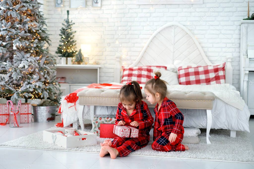 Children in pajamas unwrap gifts from Santa sitting on the floor by the bed on Christmas morning . The younger girl watches her sister untie the bow of the gift box. New Year and Xmas celebrations