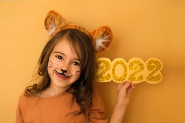 Güzel kıvırcık saçlı esmer kız ve kaplan aquagrim 'in elinde 2022' nin altın sayıları var. Kaplan kulaklı bir başlık. Yeni yıl. Kaplan kostümlü çocuk tatlı tatlı gülümser ve kameraya bakar.
