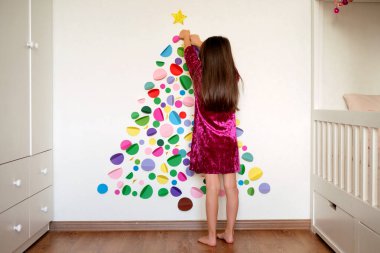 Cute girl decorates the wall in the children's room with multicolored circles forming a Christmas tree. Christmas tree made of cardboard and paper hand made. Art creative xmas tree craft. Very Peri clipart
