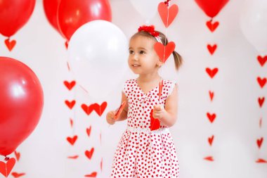 The child holds in his hand a garland with red hearts and a balloon. Bright decorations for Valentine's Day. Dress with a pattern of many hearts