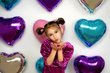 Pretty girl with ponytails sends an air kiss to all lovers, congratulating on Valentine's Day. Photo zone of bright multicolored balloons in the shape of a hearts