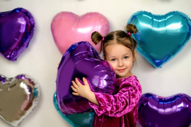 A cute three-year-old girl hugs a purple foil balloon in the shape of a heart. Decor photo zone of hearts for Valentine's Day