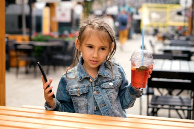 Elinde bir bardak limonata ve elinde akıllı telefon olan bir çocuk düşünceli hayalperest bir bakışla yana bakıyor. Güneşli bir yaz gününde popüler bir şehir kafesinin önünde bir kız oturuyor.