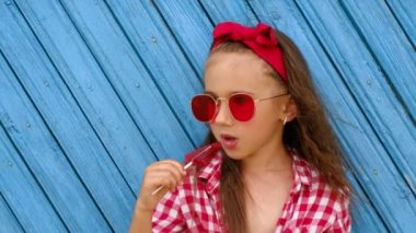 Kırmızı güneş gözlüklü kız elinde çubukla üçgen bir lolipop tutuyor, diliyle yalıyor. Mavi panoda şeker yiyen bir çocuğun profili