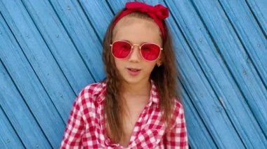 Kız elleri ağzında yüksek sesle haber bağırıyor. Kırmızı güneş gözlüklü, mavi arka planda ekose gömlekli sevimli bir anaokulu öğrencisi. Bebek yüzünün duygusal ifadesi.