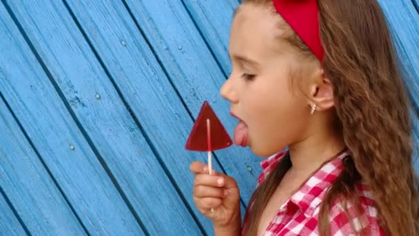 頭に赤い弓を持つ美しい長髪の女の子は プロフィールで横に立って 大きなロリポップをなめる お菓子の害や大量の砂糖の使用 明るいジューシーな組み合わせ — ストック動画
