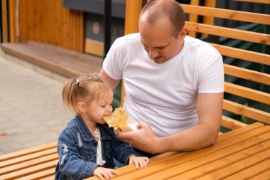 Bir baba her zaman kızıyla ilgilenir, karnını doyurur ve onu gücendirmesine izin vermez. Komik küçük bir kız lezzetli bir öğle yemeğinden bir ısırık alıyor. Babası beyaz tişörtlü, kızı kot ceketli.