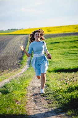 Sahada elinde hasır şapkayla koşan mutlu kıvırcık esmer. Mavi elbiseli genç bir kadın doğada yürüyor. Yeşil bahar çayırı.