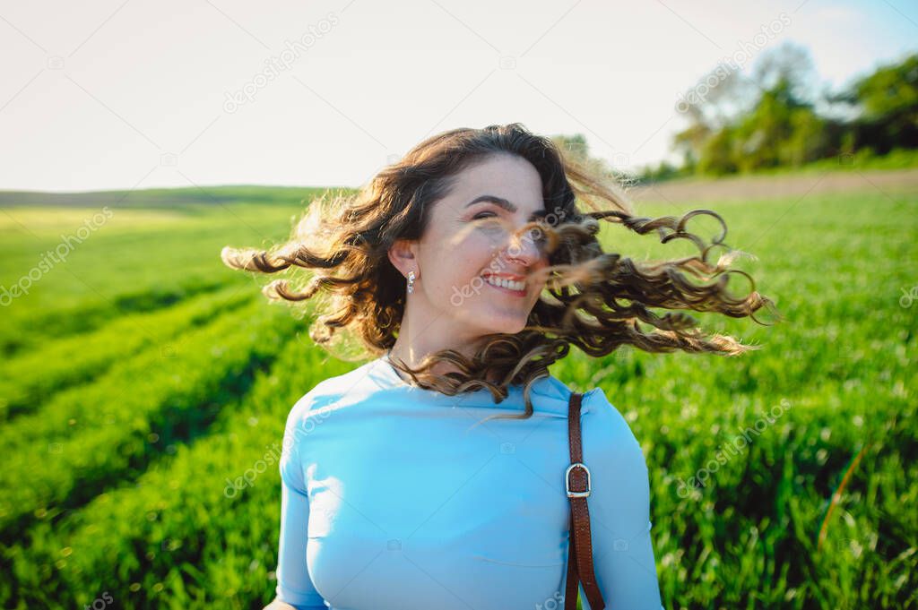 Curly brunette enjoys sunshine in summer day. Portrait of smiling beautiful girl outdoors. Young woman standing on street with curly hair blown by the wind, covering her face.