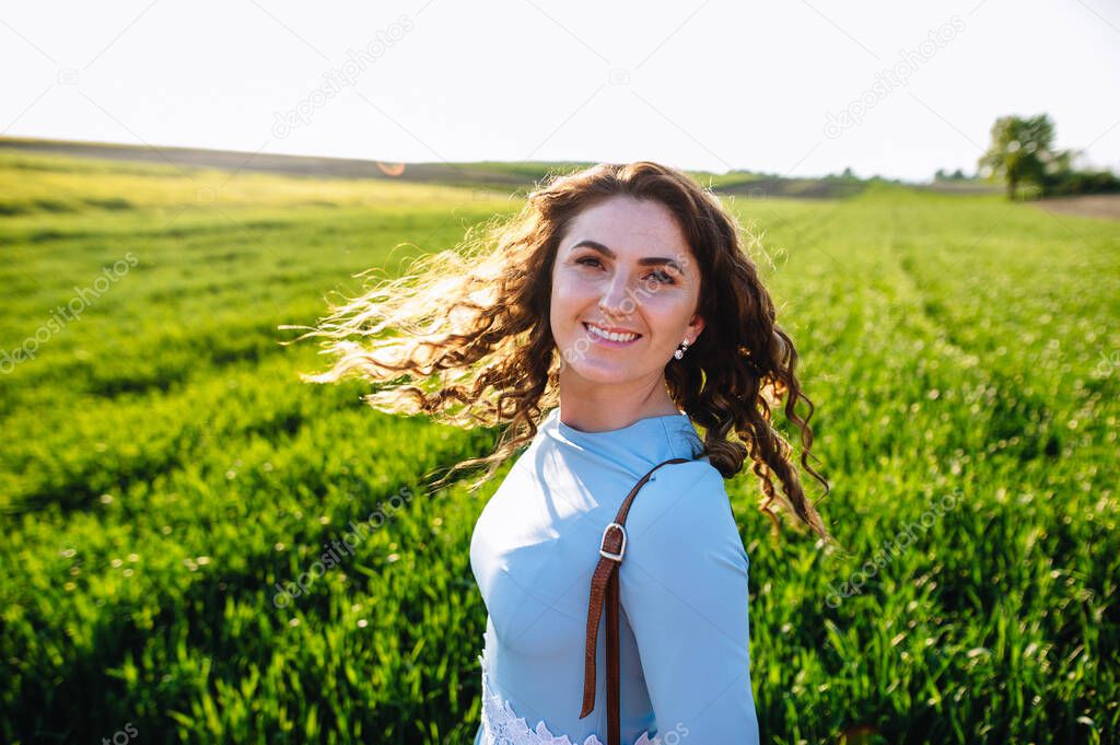 Curly brunette enjoys sunshine in summer day. Portrait of smiling beautiful girl outdoors. Young woman standing on street with curly hair blown by the wind, covering her face.