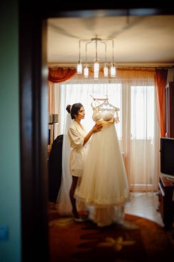 Beyaz bir pignoir giymiş güzel bir gelin, gelinliğinin yanında duruyor ve gelinlik askıda asılı duruyor. Sabah Gelini Toplantısı