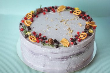 Yaban mersini, kızılcık, portakal ve koni ile süslenmiş menekşe kremalı pasta. Mavi arka planda gri yuvarlak pasta, yeni yıl tarzında dekore edilmiş.