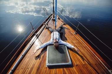 Güneş gözlüklü yatçı yatın burnuna bakıp gülüyor. Güneş suyun içinde parlıyor.
