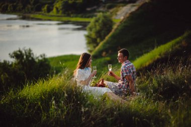 Nehir kıyısında gün batımında şarap içen sevgi dolu bir çift. Bir erkek arkadaş ve bir kız romantik bir randevuda, meyve sepetinin yanında oturup bardaktan şampanya içiyorlar..