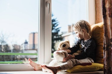 Pencerede otururken gün ışığının tadını çıkaran tatlı küçük kız. Çocuk evde otururken pencereden dışarı bakar. D vitamini