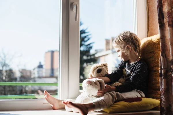 Cute little girl enjoying the sunshine while sitting at the window. The child looks out the window while sitting in the house. Vitamin D