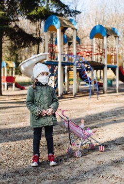 Kapalı oyun alanının arka planında koruyucu maskeli üzgün küçük kız. Tıbbi maskeli bir çocuk parkta yürüyor. COVID-19 'a karşı karantina önlemleri. Koronavirüs salgını.