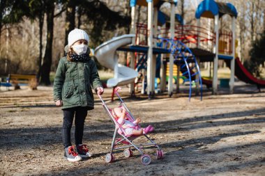 Kapalı oyun alanının arka planında koruyucu maskeli üzgün küçük kız. Tıbbi maskeli bir çocuk parkta yürüyor. COVID-19 'a karşı karantina önlemleri. Koronavirüs salgını.