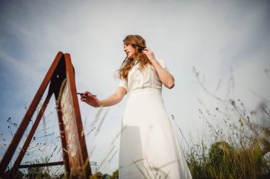 Uzun kıvırcık saçlı güzel kız elinde fırçayla sehpanın yanında duruyor. Ressam kız açık havada resim yapıyor..