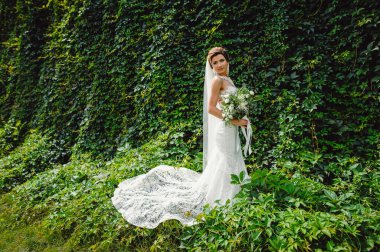 Lüks bir gelinlik içindeki güzel gelin açık havada bir buket beyaz gül ve yeşillik tutuyor. Beyaz elbiseli mutlu gelinin portresi duvarın arkasında yeşilliklerle gülümsüyor..