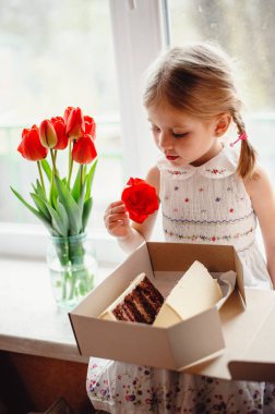 Dört yaşında güzel bir kız, bir odanın pencere eşiğinde oturur ve elinde çikolatalı pastayla bir kutu tutar. Camda bir buket kırmızı lale var. Beyaz işlemeli elbiseli sevimli sarışın.