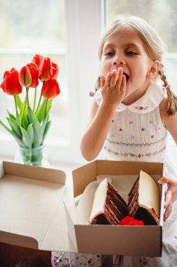 Dört yaşında güzel bir kız, bir odanın pencere eşiğinde oturur ve elinde çikolatalı pastayla bir kutu tutar. Camda bir buket kırmızı lale var. Beyaz işlemeli elbiseli sevimli sarışın.