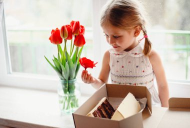 Dört yaşında güzel bir kız, bir odanın pencere eşiğinde oturur ve elinde çikolatalı pastayla bir kutu tutar. Camda bir buket kırmızı lale var. Beyaz işlemeli elbiseli sevimli sarışın.