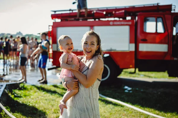 Happy mother holds a little daughter in her arms that cries. Wet mom with her daughter, on the background of a fire truck.