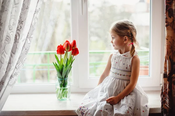 一个四岁的漂亮小女孩 坐在房间的窗台上 喜庆地捧着一束红郁金香 漂亮的金发碧眼的白色绣花衣服 快乐的孩子 免版税图库照片