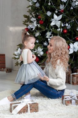 Beyaz kazaklı yakışıklı bir anne süslü bir Noel ağacının arka planında küçük kızıyla oturuyor. Birbirlerine bakıyorlar. Portreler. Kopyalama alanı.