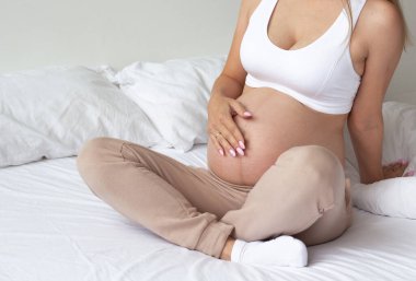 Evdeki yatakta oturan tatlı sarışın hamile kız. Hafif bir arkaplanda. Portre. 8 aylık hamilelik. Bir bebeğin doğmasını bekleme konsepti. Karnını yaklaştır. Kopyalama alanı.