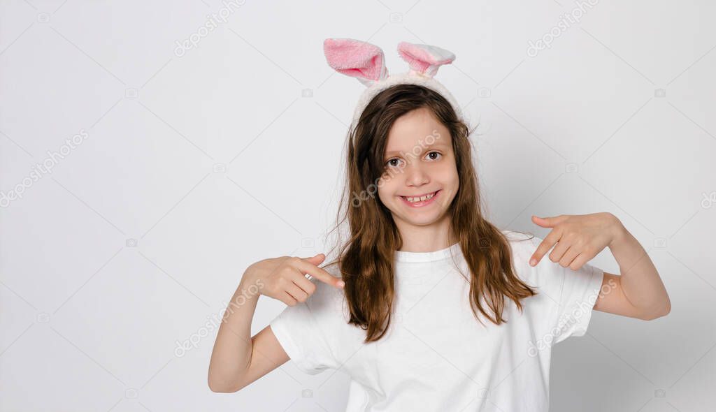 Cute dark-haired girl 9 years old. With bunny ears on the head. Pointing at a white T-shirt. Easter Bunny. close-up. High quality photo. copy space. Panarama 