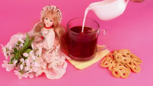 Milk pouring in tea in glass mug and doll in pink lighting on pink background. — Stockvideo