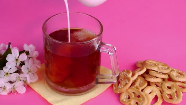 Milk pouring in tea in glass mug and cookies and pretzels, in pink lighting on pink background. — Wideo stockowe