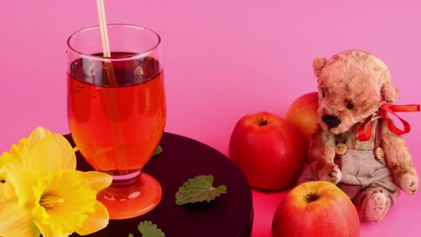 Apple juice or cider in transparent drinking glass rotate slowly on turntable in pink light. Teddy bear and three fresh apple near. — Video