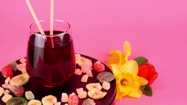 Fruktjuice eller cocktail i genomskinligt dricksglas roterar långsamt på skivtallriken i rosa ljus. Olika torkade frukter och vårblommor nära. — Stockvideo