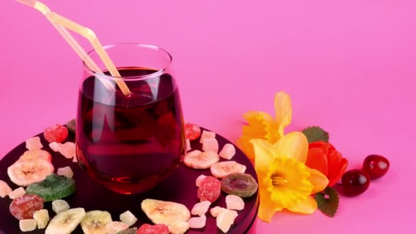 Fruit juice or cocktail in transparent drinking glass rotate slowly on turntable in pink light. Different dried fruits and spring flowers near. — Stok video