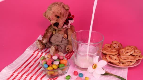 在椒盐饼干、糖果和泰迪熊旁边的杯子里倒入新鲜的农场牛奶。粉红的灯光. — 图库视频影像