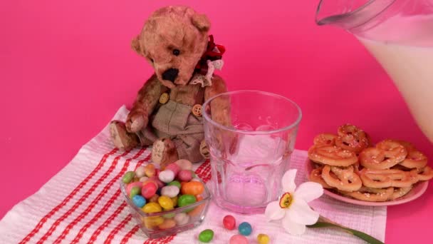 Frische Bauernmilch neben Brezeln, Bonbons und Teddybär ins Glas gießen. Mit rosa Licht. — Stockvideo